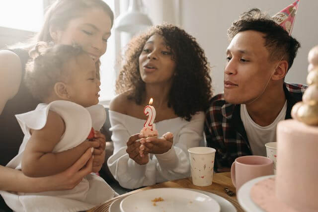 adults celebrating 2-year-old's birthday with lit candle.